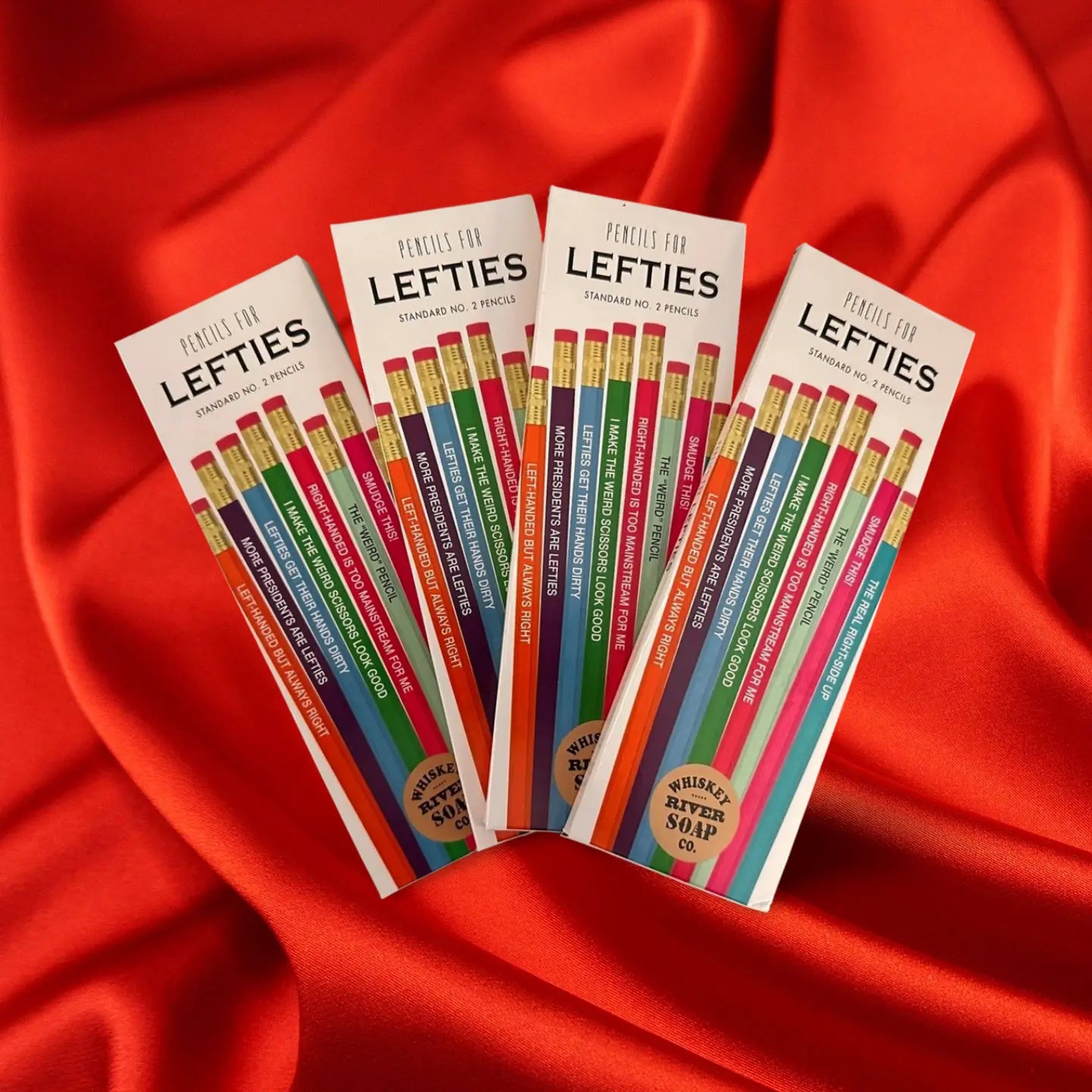 Four packs of “Pencils for Lefties” displayed on a wooden table, featuring humorous left-handed themed No. 2 pencils in assorted colors.