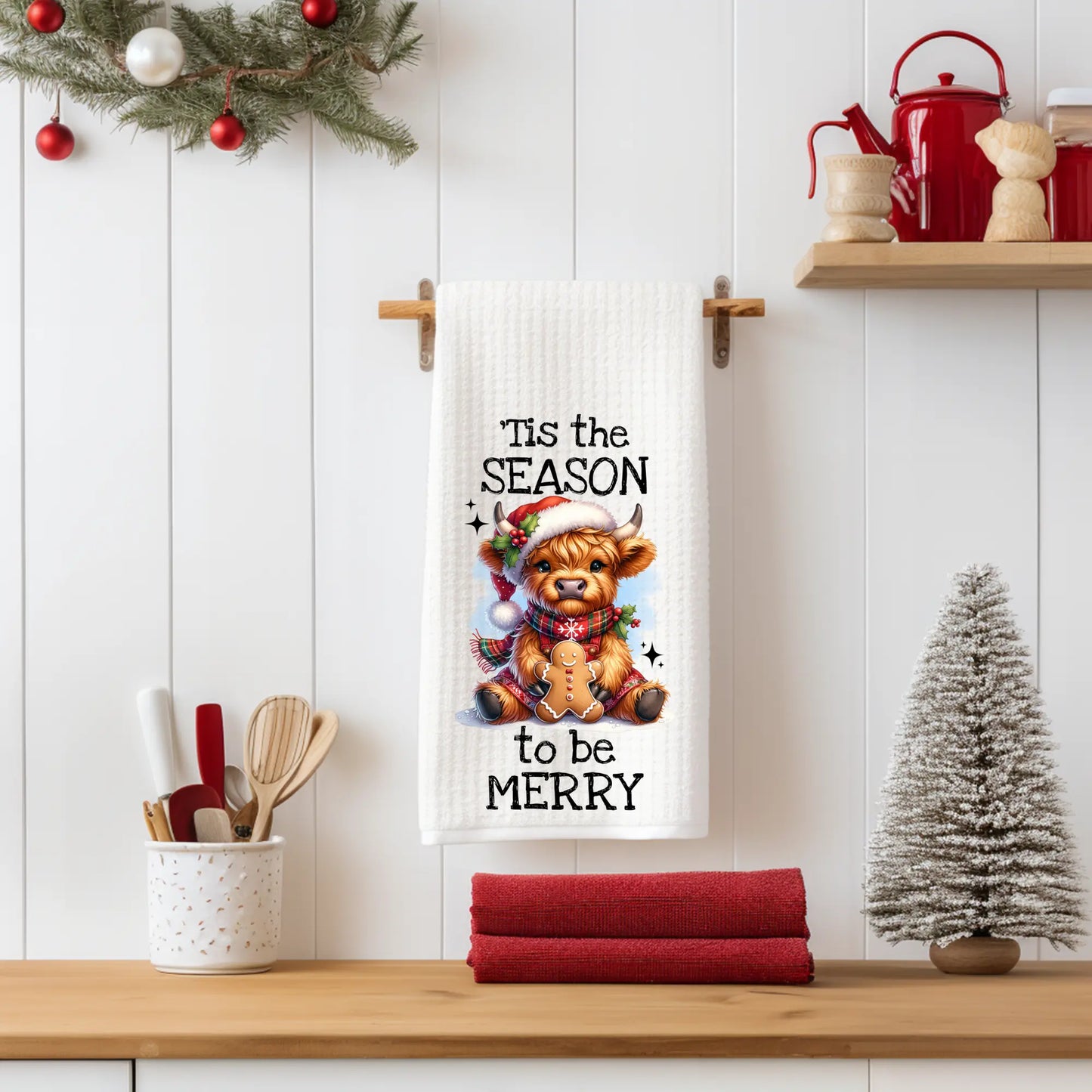 White 18x28 polyester waffle-weave tea towel featuring a festive Highland cow holding a gingerbread cookie with the text “’Tis the Season to Be Merry.” Displayed in a holiday kitchen scene with utensils, red décor, and a miniature Christmas tree.