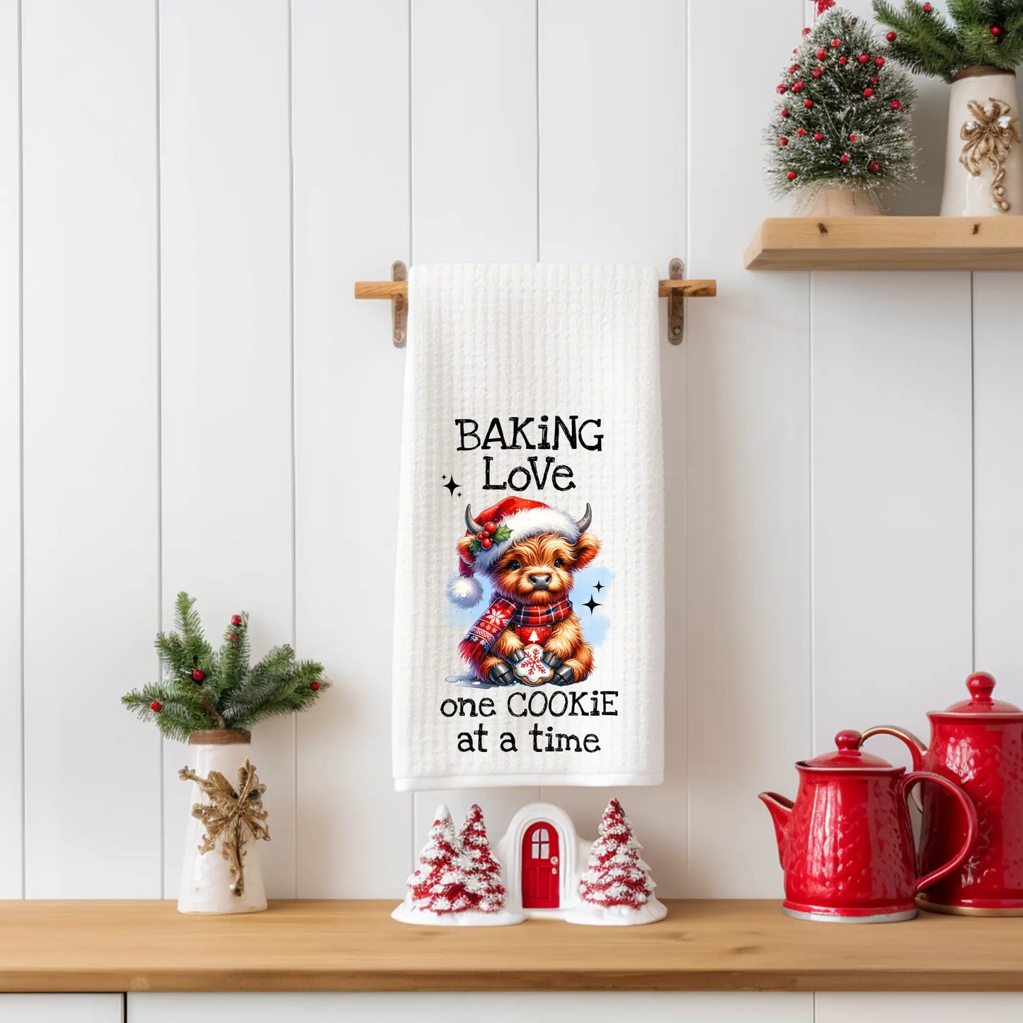 White 18x28 polyester waffle-weave tea towel hanging on a wooden peg rail, featuring a festive Highland cow holding a cookie with the text “Baking Love One Cookie at a Time.” Displayed in a holiday kitchen scene with red teapots, mini Christmas trees, and seasonal décor.