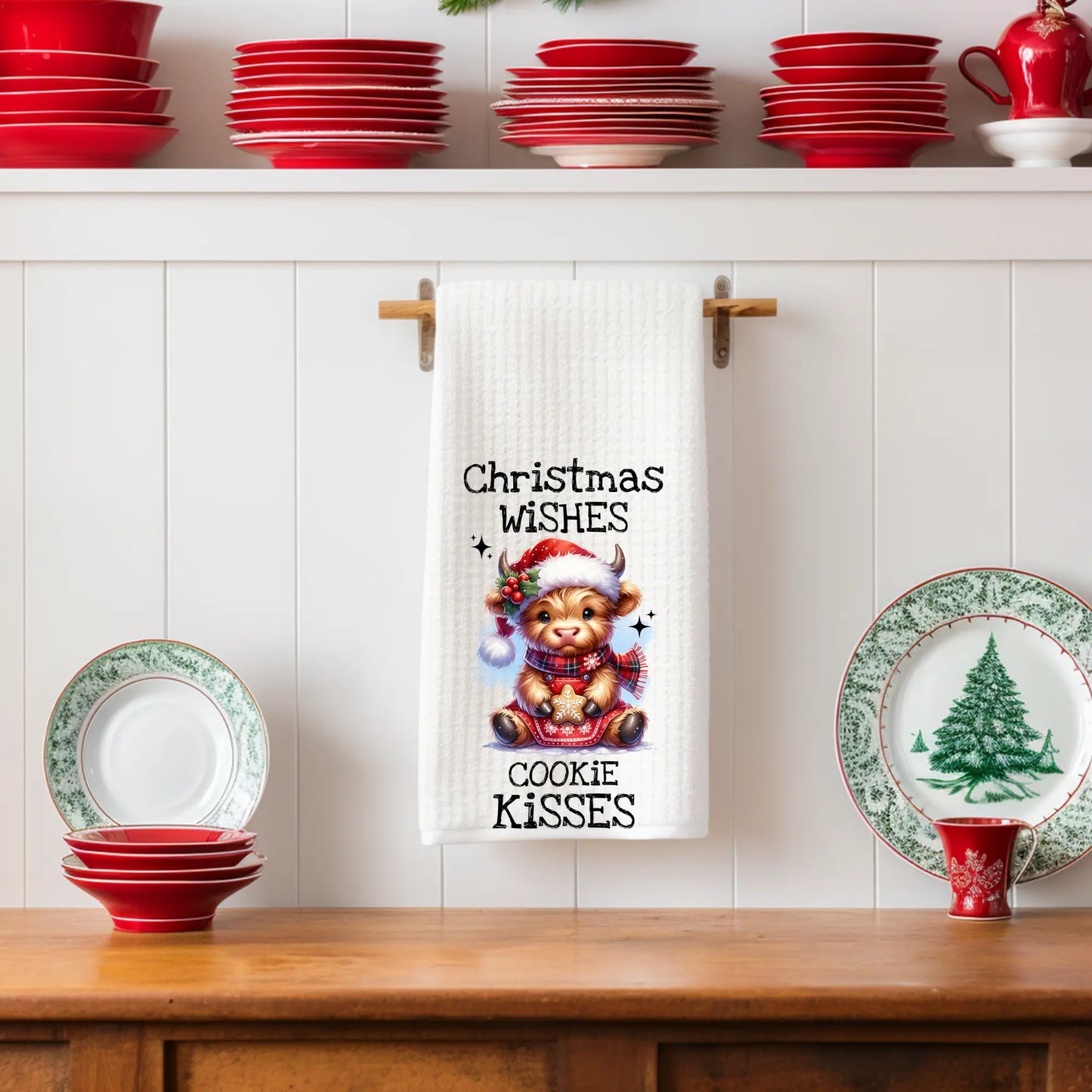White 18x28 polyester waffle-weave tea towel hanging on a wooden peg rail, featuring a festive Highland cow holding a cookie with the text “Christmas Wishes Cookie Kisses.” Displayed in a holiday kitchen scene with red dishes, Christmas plates, and decorative shelving.