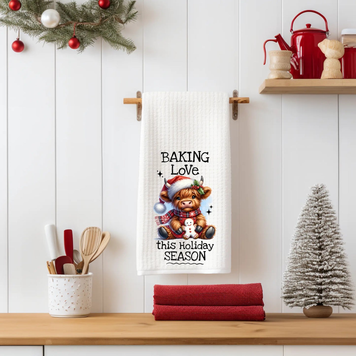 White 18x28 polyester waffle-weave tea towel hanging on a wooden peg rail, featuring a festive Highland cow holding a gingerbread cookie with the text “Baking Love This Holiday Season.” Displayed in a holiday kitchen scene with utensils, pinecones, a mini Christmas tree, and red décor.