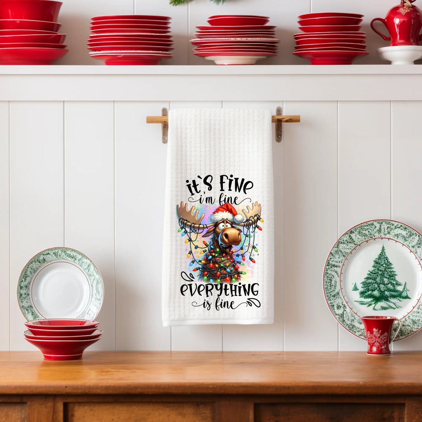 White 18x28 polyester waffle-weave Christmas tea towel hanging on a wooden peg rail, featuring a festive moose wearing a Santa hat and wrapped in colorful Christmas lights with the text “’Tis the Season to Be Jolly.” Displayed in a holiday kitchen scene with small Christmas trees, red teapots, and winter décor.