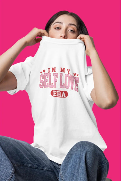 Model wearing a white t-shirt reading “In my self love era” with pink Valentine-style lettering on a bright pink background