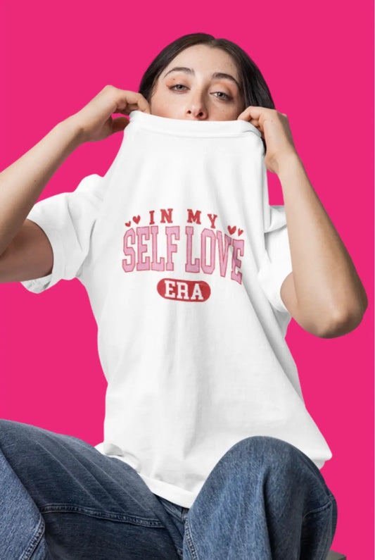Model wearing a white t-shirt reading “In my self love era” with pink Valentine-style lettering on a bright pink background