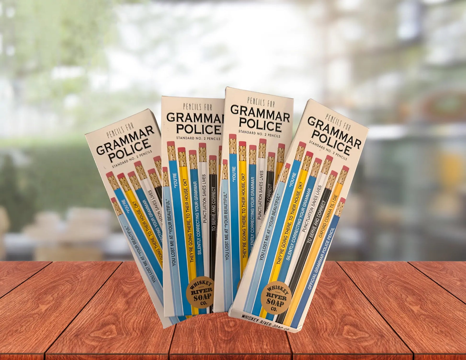 Four packs of “Pencils for Grammar Police” displayed on a wooden table, featuring blue and yellow No. 2 pencils printed with funny grammar quotes.
