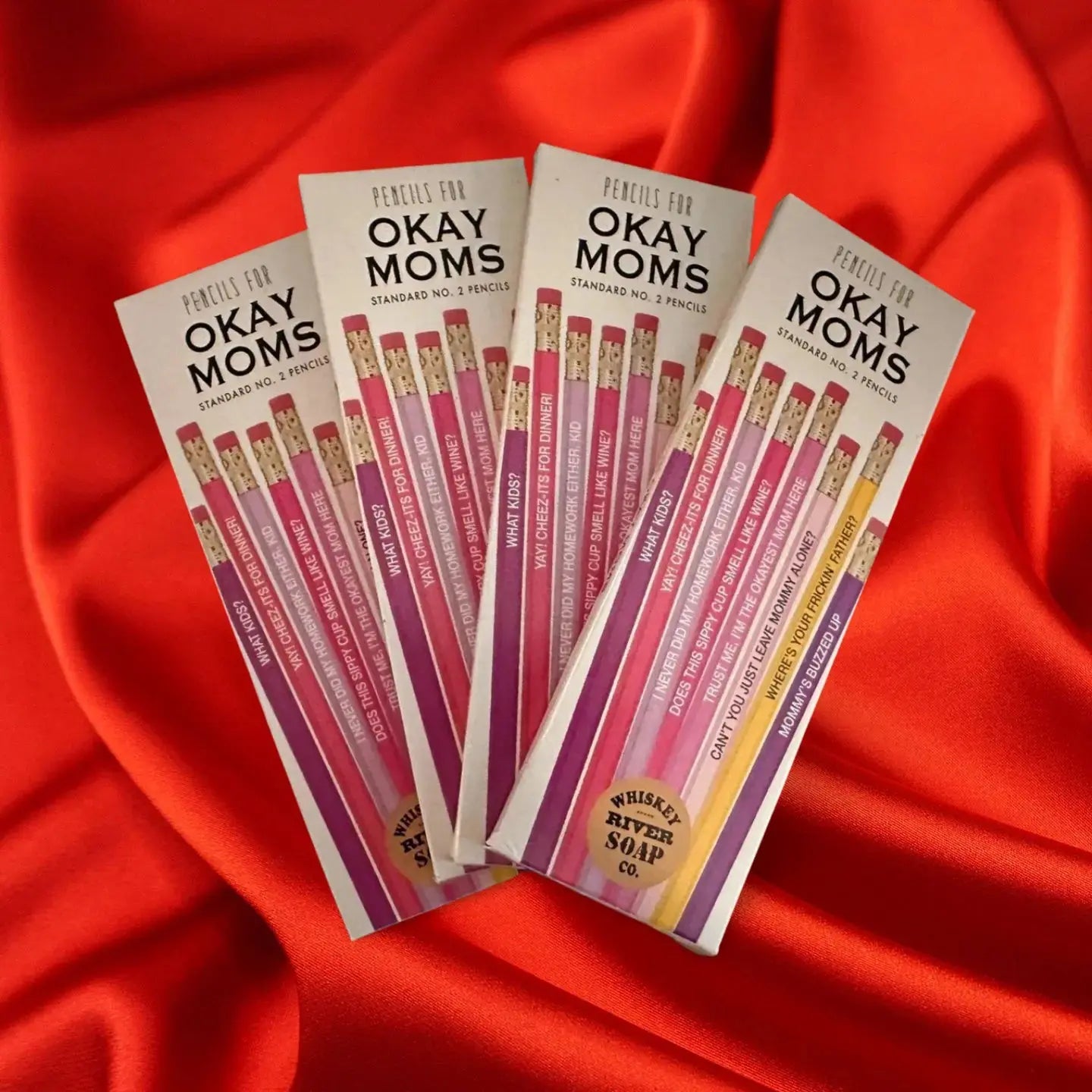 Four packs of “Pencils for Okay Moms” displayed on a wooden table, featuring pink No. 2 pencils printed with funny motherhood sayings.