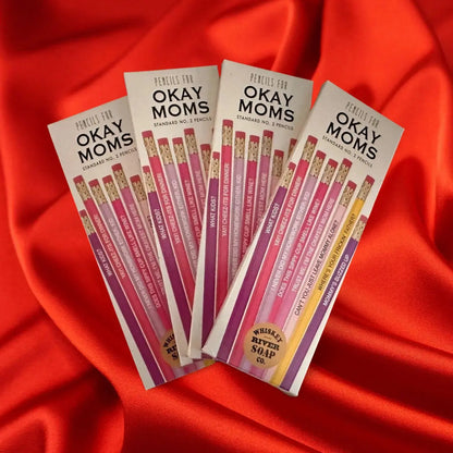 Four packs of “Pencils for Okay Moms” displayed on a wooden table, featuring pink No. 2 pencils printed with funny motherhood sayings.