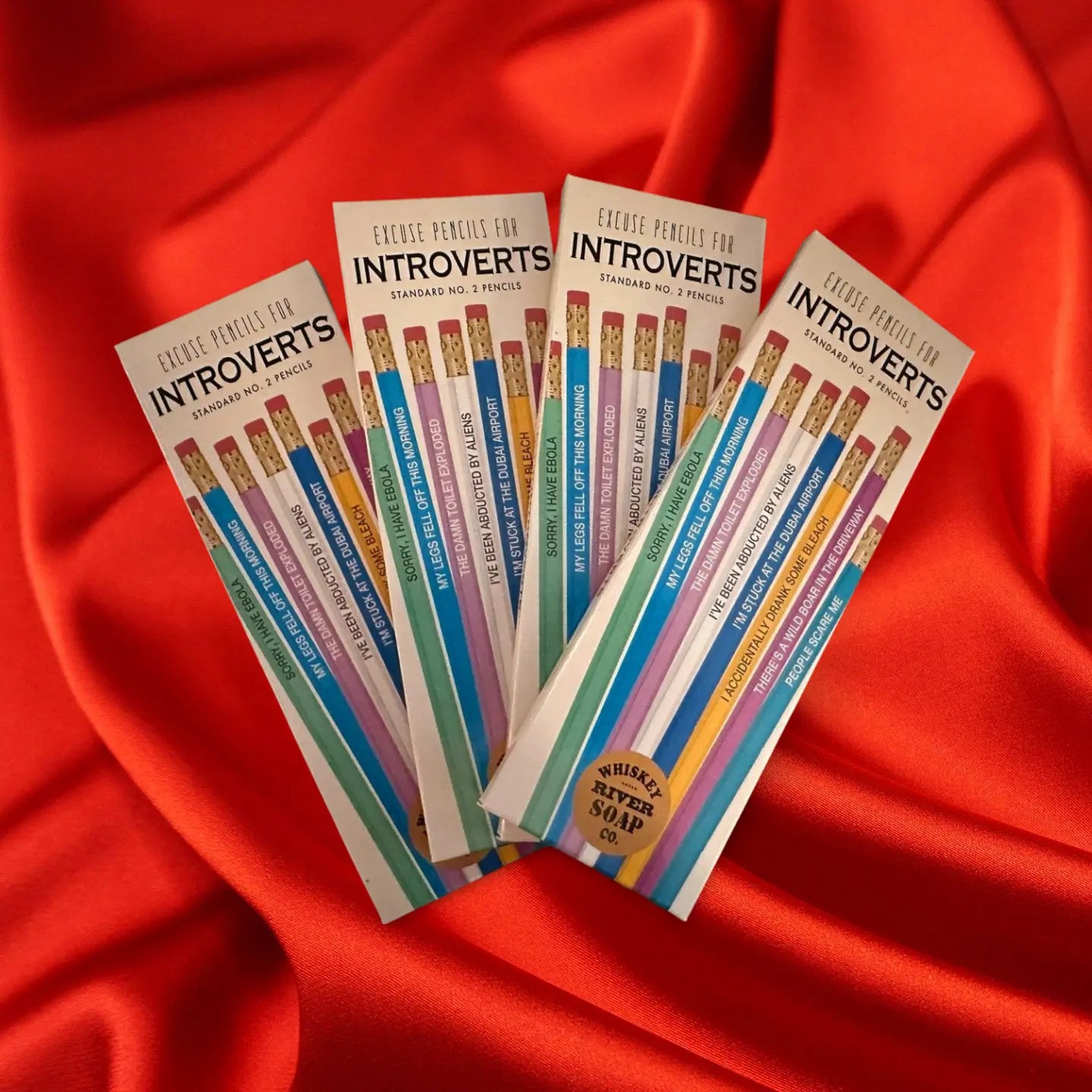 Four packs of “Pencils for Okay Moms” displayed on a wooden table, featuring pink No. 2 pencils printed with funny motherhood sayings.