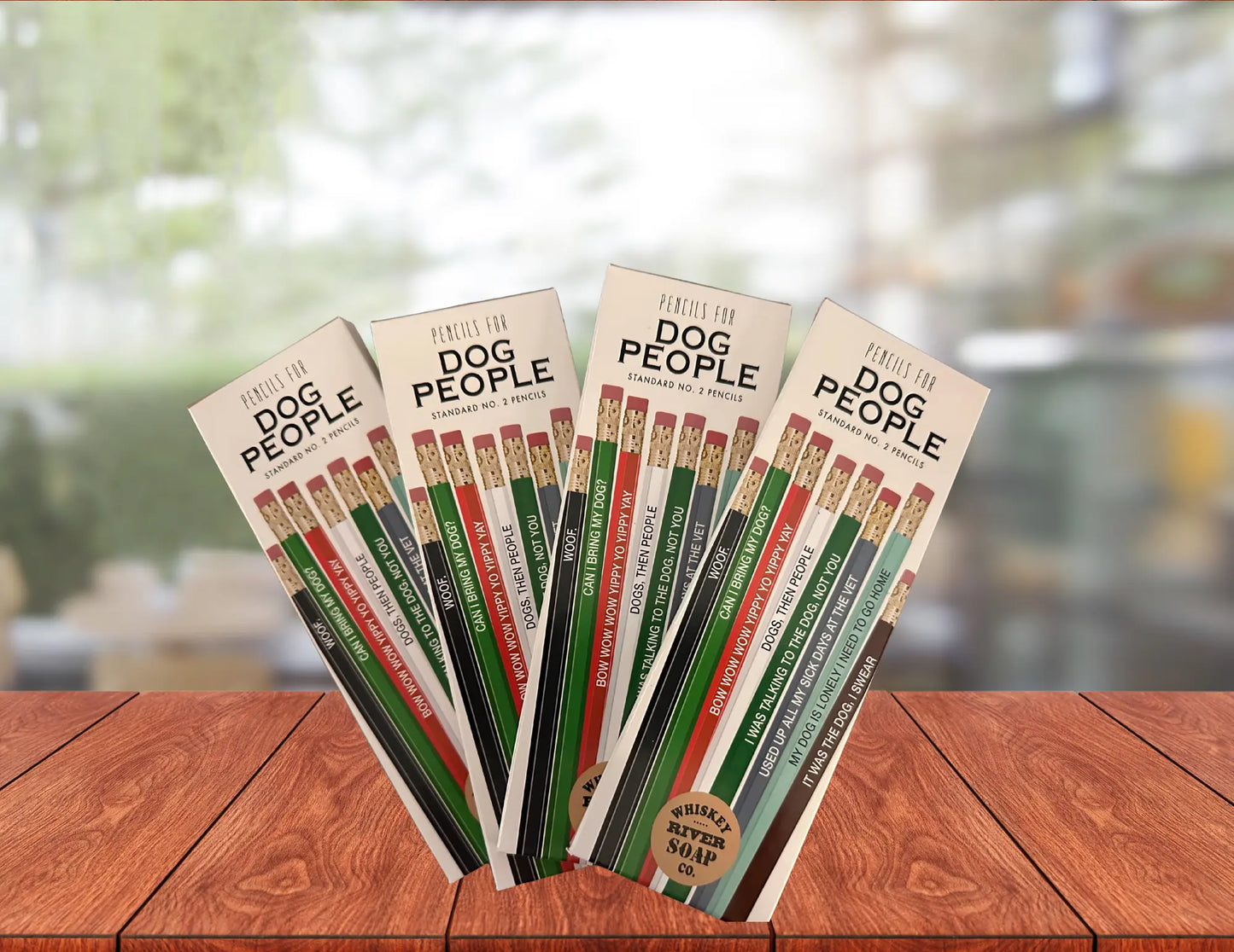 Packaged sets of “Pencils for Dog People” displayed upright on a wooden table, featuring colorful novelty No. 2 pencils with humorous dog-lover phrases.