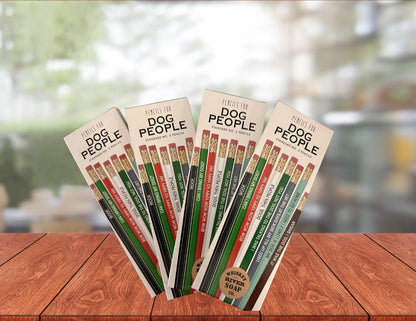 Packaged sets of “Pencils for Dog People” displayed upright on a wooden table, featuring colorful novelty No. 2 pencils with humorous dog-lover phrases.