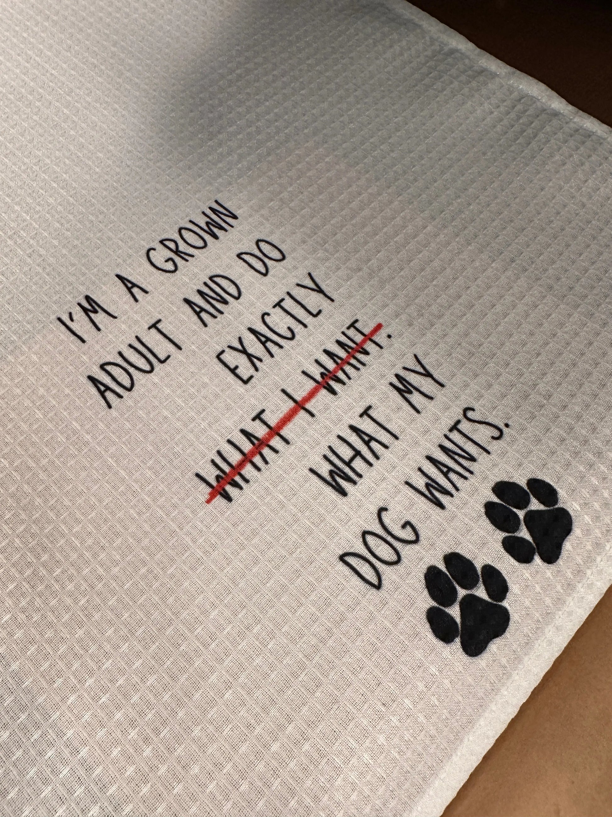 White 18x28 polyester waffle-weave tea towel with the phrase “I’m a grown adult and do exactly what my dog wants,” featuring crossed-out text and two black paw prints.