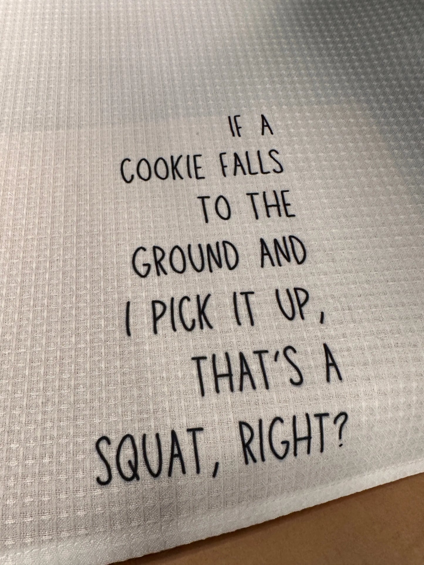 Close-up of a white 18x28 polyester waffle-weave tea towel printed with the phrase “If a cookie falls to the ground and I pick it up, that’s a squat, right?” in black lettering.