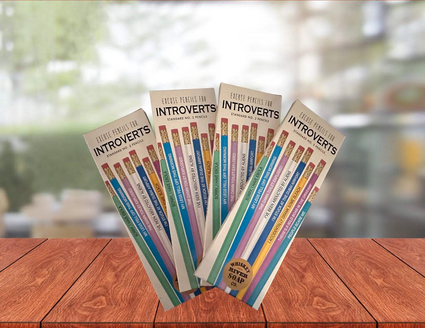 Four packs of “Pencils for Okay Moms” displayed on a wooden table, featuring pink No. 2 pencils printed with funny motherhood sayings.