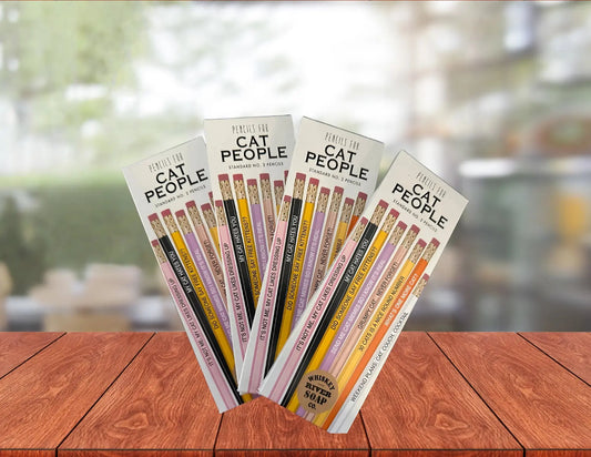 Four packs of “Pencils for Okay Moms” displayed on a wooden table, featuring pink No. 2 pencils printed with funny motherhood sayings.