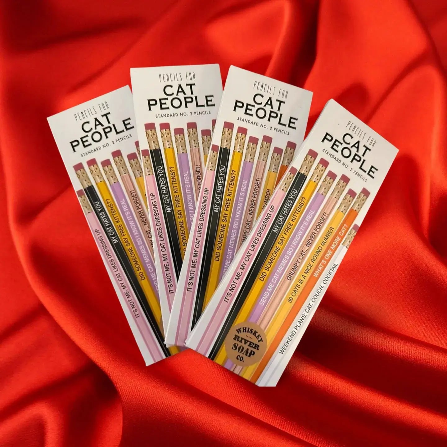 Four packs of “Pencils for Okay Moms” displayed on a wooden table, featuring pink No. 2 pencils printed with funny motherhood sayings.
