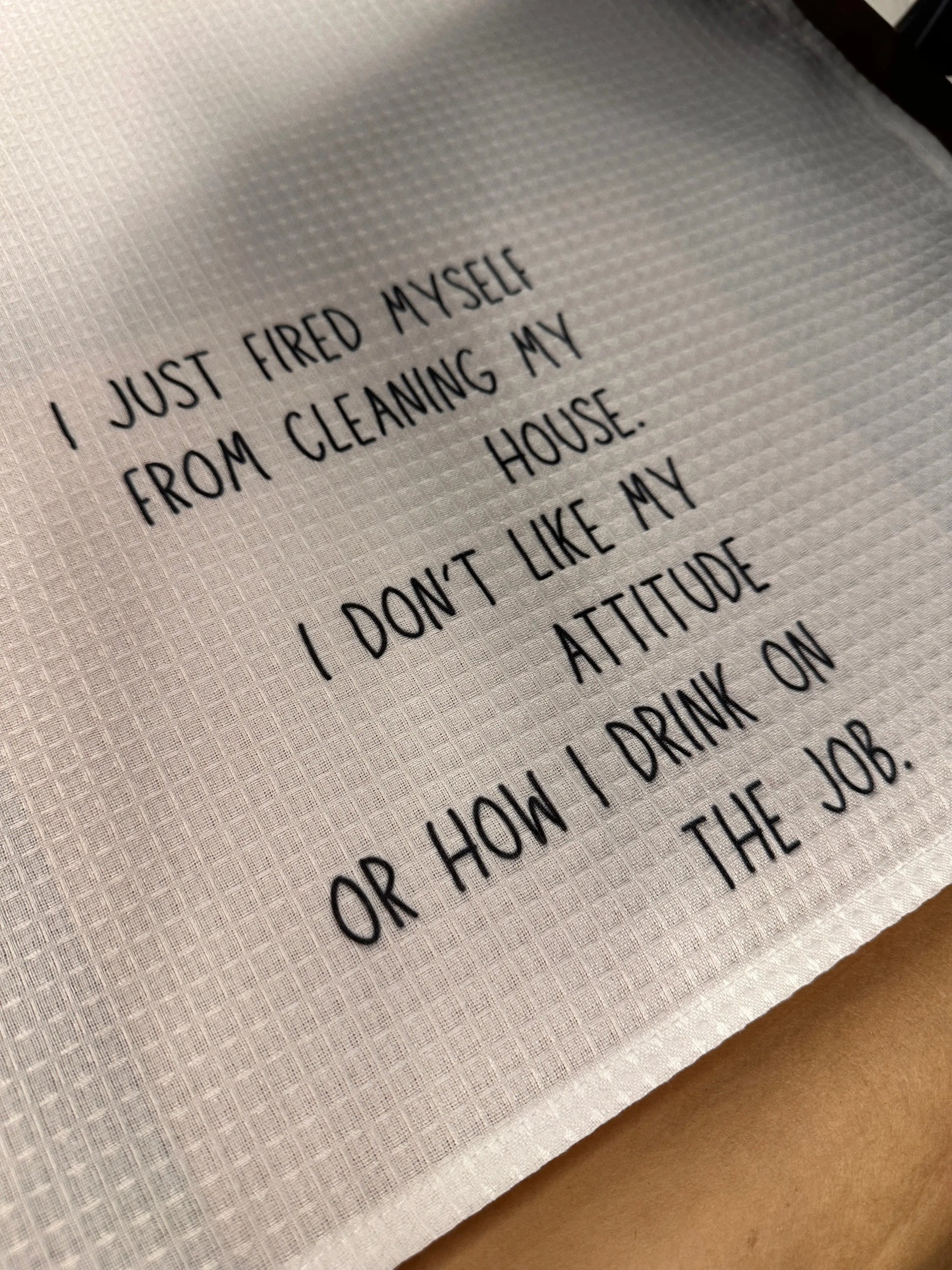 Close-up of a white 18x28 polyester waffle-weave tea towel printed with the phrase “I just fired myself from cleaning my house. I don’t like my attitude or how I drink on the job.” in black lettering.