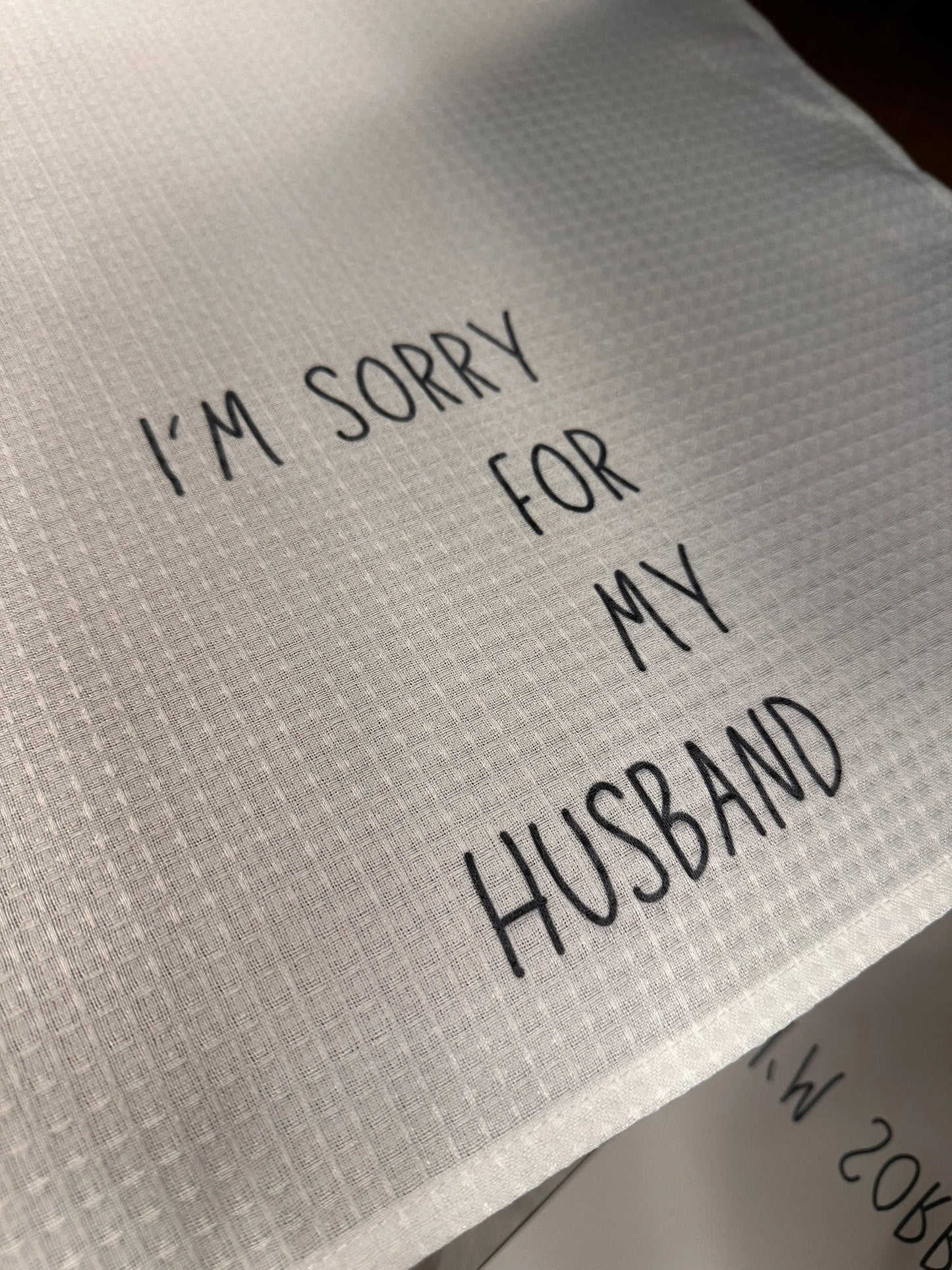 White waffle-weave 18x28 tea towel printed with the phrase “I’m sorry for my husband” in black lettering.
