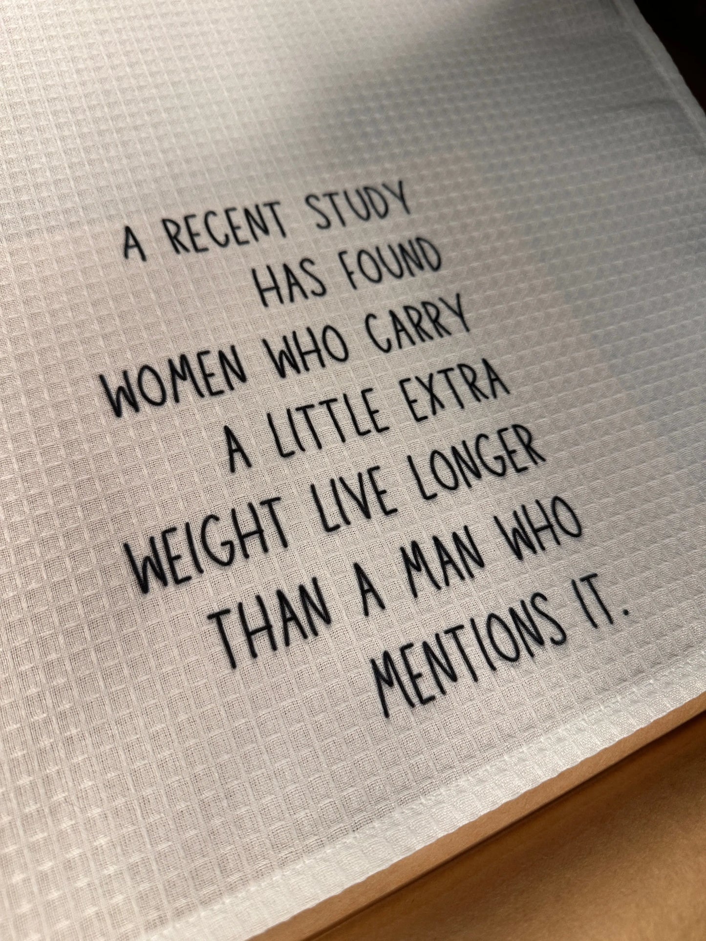 White 18x28 polyester waffle-weave tea towel printed with the humorous phrase “A recent study has found women who carry a little extra weight live longer than a man who mentions it.” in black lettering.