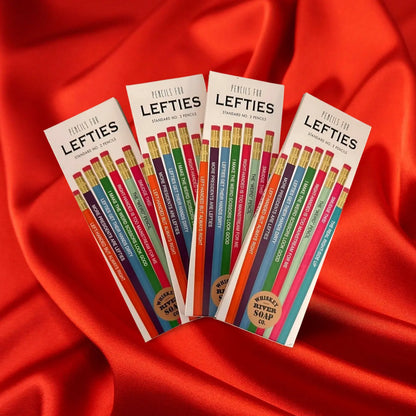 Four packs of “Pencils for Lefties” displayed on a wooden table, featuring humorous left-handed themed No. 2 pencils in assorted colors.