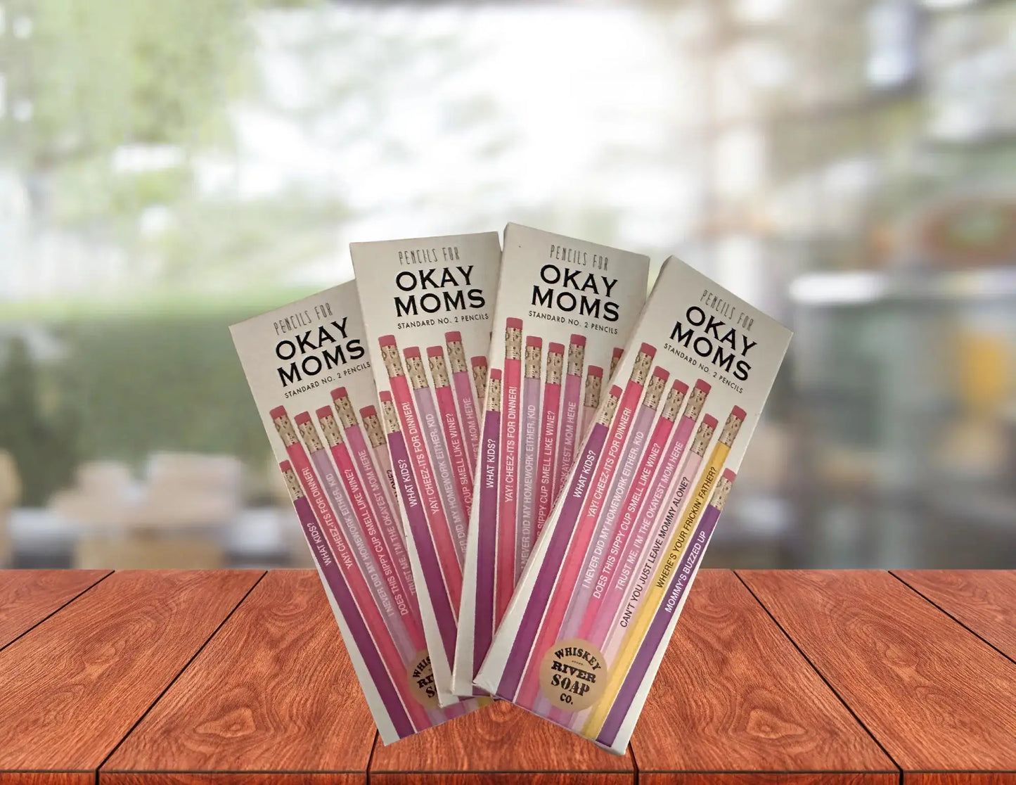 Four packs of “Pencils for Okay Moms” displayed on a wooden table, featuring pink No. 2 pencils printed with funny motherhood sayings.