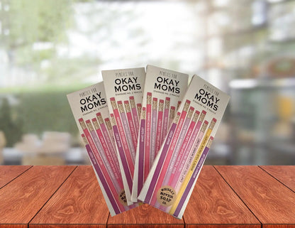 Four packs of “Pencils for Okay Moms” displayed on a wooden table, featuring pink No. 2 pencils printed with funny motherhood sayings.
