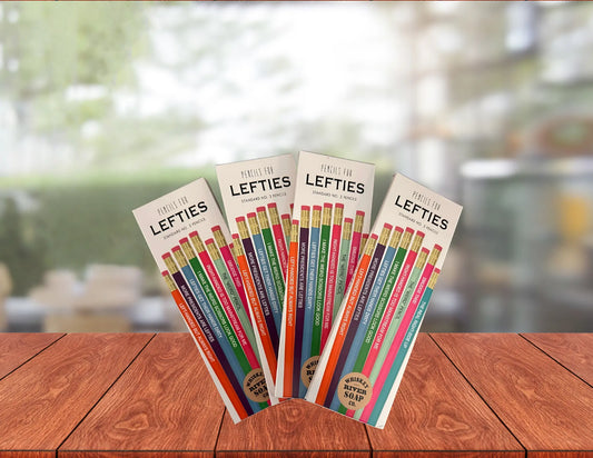Four packs of “Pencils for Lefties” displayed on a wooden table, featuring humorous left-handed themed No. 2 pencils in assorted colors.