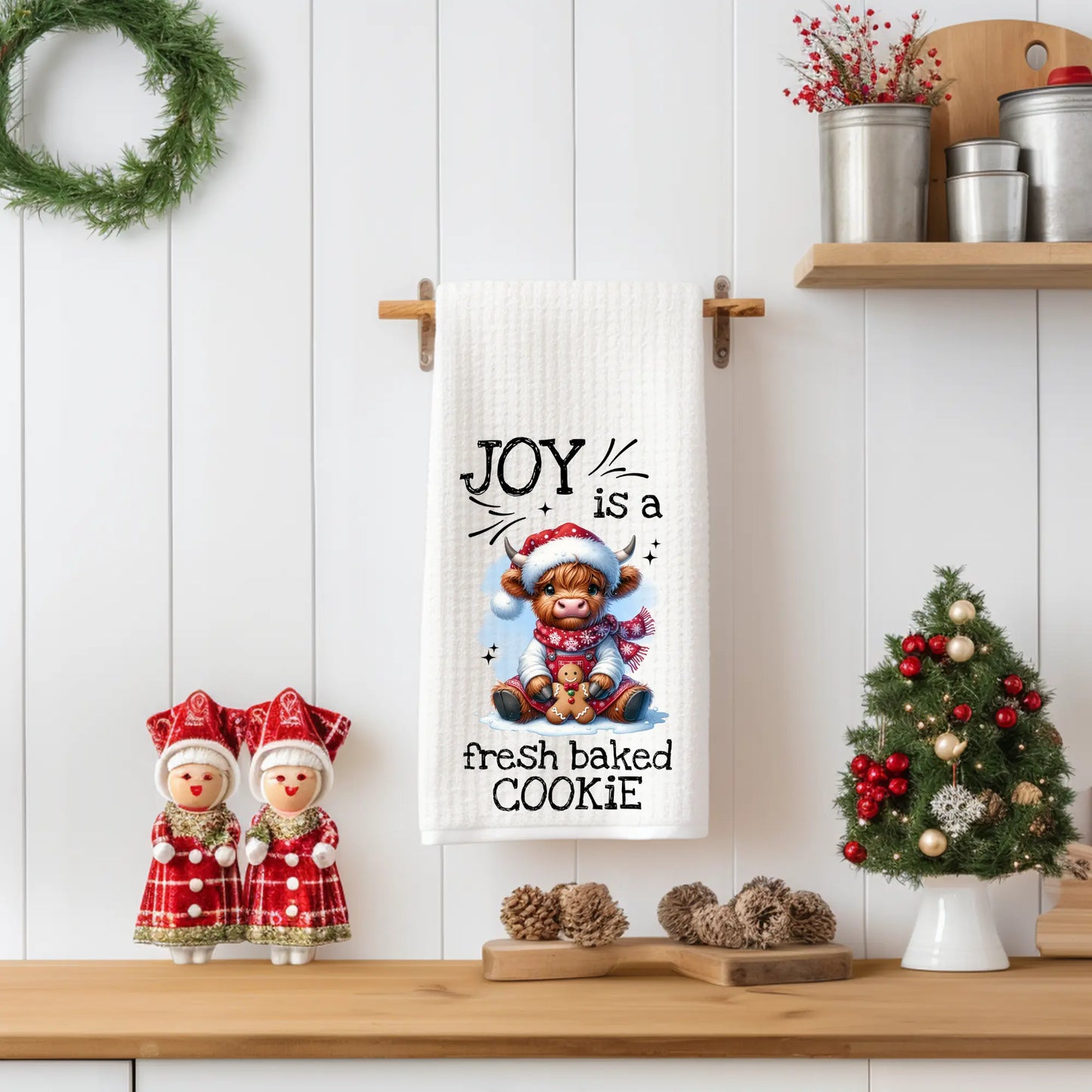 White 18x28 polyester waffle-weave tea towel featuring a festive Highland cow holding a cookie with the text “Joy Is a Fresh Baked Cookie.” Displayed in a holiday kitchen scene with red décor, pinecones, and a miniature Christmas tree.