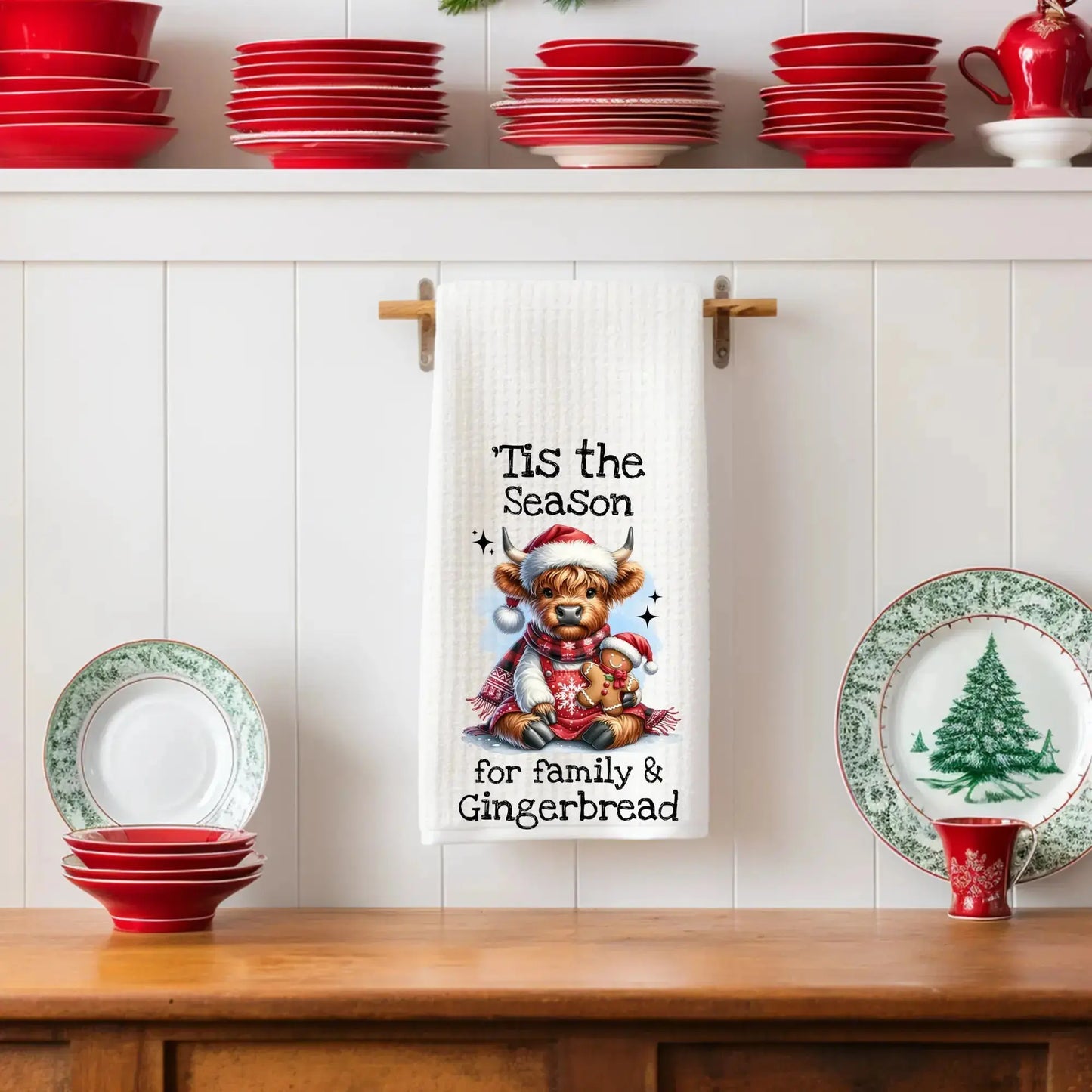 White 18x28 polyester waffle-weave tea towel featuring a festive Highland cow holding a gingerbread cookie with the text “’Tis the Season for Family & Gingerbread.” Displayed in a holiday kitchen scene with red dishes and Christmas plates.