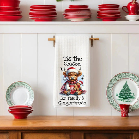 White 18x28 polyester waffle-weave tea towel featuring a festive Highland cow holding a gingerbread cookie with the text “’Tis the Season for Family & Gingerbread.” Displayed in a holiday kitchen scene with red dishes and Christmas plates.