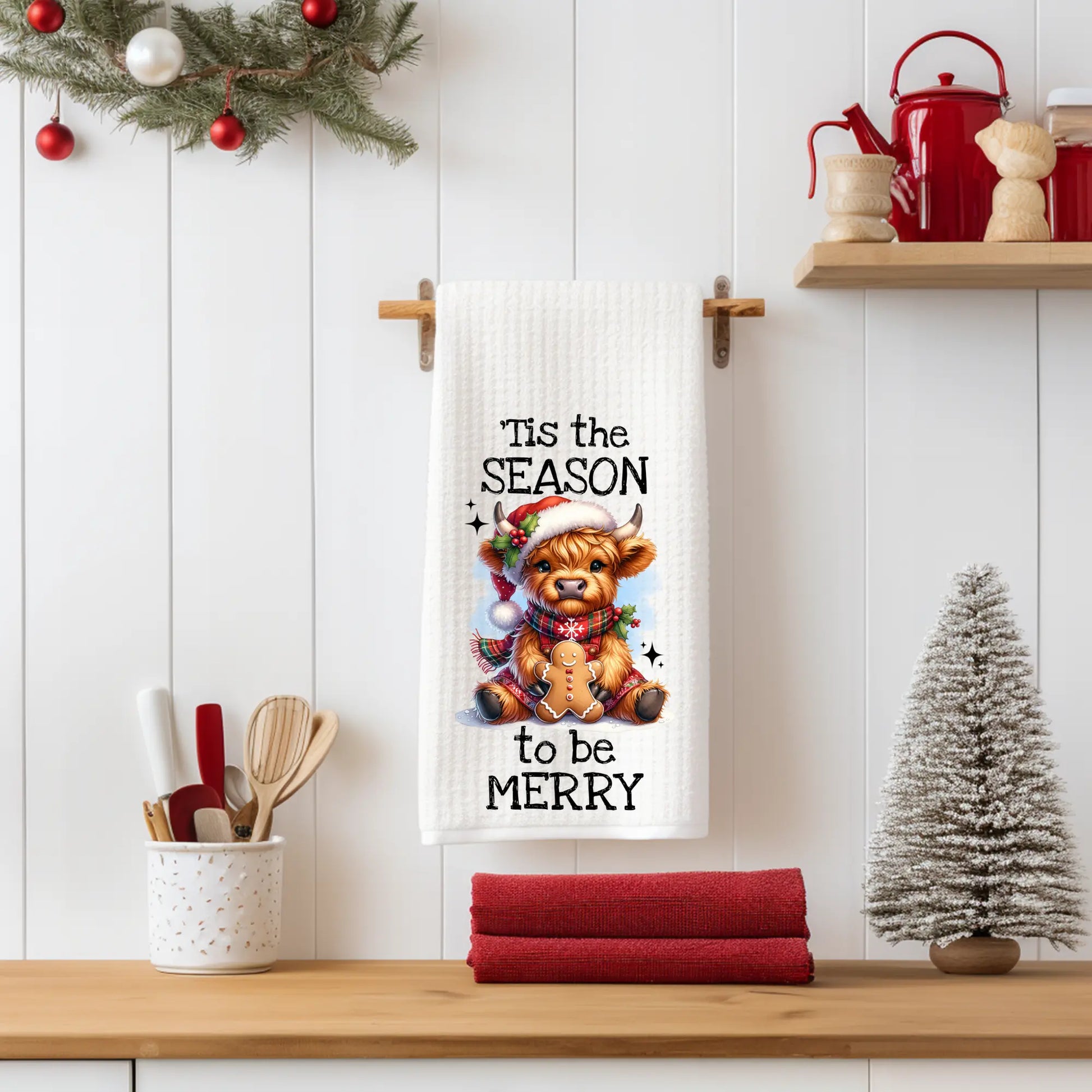 White 18x28 polyester waffle-weave tea towel featuring a festive Highland cow holding a gingerbread cookie with the text “’Tis the Season to Be Merry.” Displayed in a holiday kitchen scene with utensils, red décor, and a miniature Christmas tree.