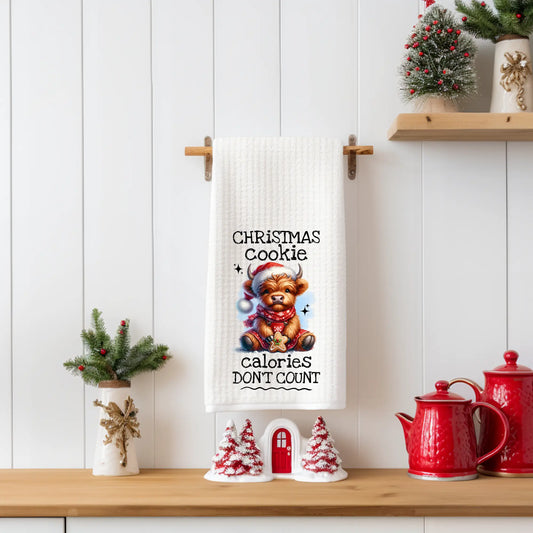White 18x28 polyester waffle-weave tea towel featuring a festive Highland cow holding a cookie with the text “Christmas Cookie Calories Don’t Count.” Displayed in a holiday kitchen scene with red teapots, mini Christmas trees, and seasonal décor.