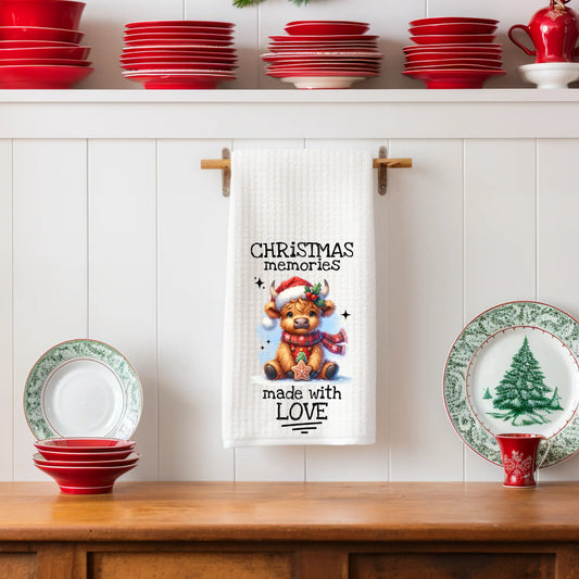 White 18x28 polyester waffle-weave tea towel hanging on a wooden peg rail, featuring a festive Highland cow holding a cookie with the text “Christmas Memories Made With Love.” Displayed in a holiday kitchen scene with red dishes, Christmas plates, and pinecone décor.