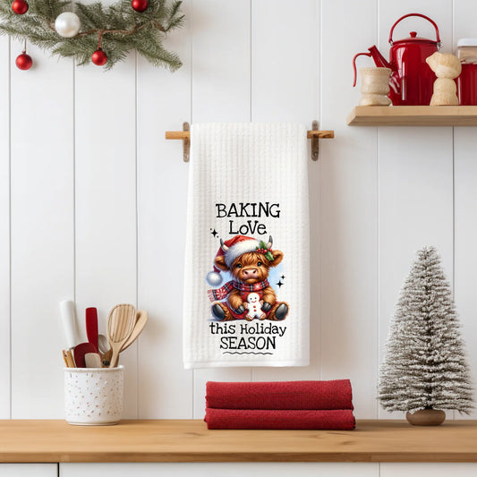 White 18x28 polyester waffle-weave tea towel hanging on a wooden peg rail, featuring a festive Highland cow holding a gingerbread cookie with the text “Baking Love This Holiday Season.” Displayed in a holiday kitchen scene with utensils, pinecones, a mini Christmas tree, and red décor.