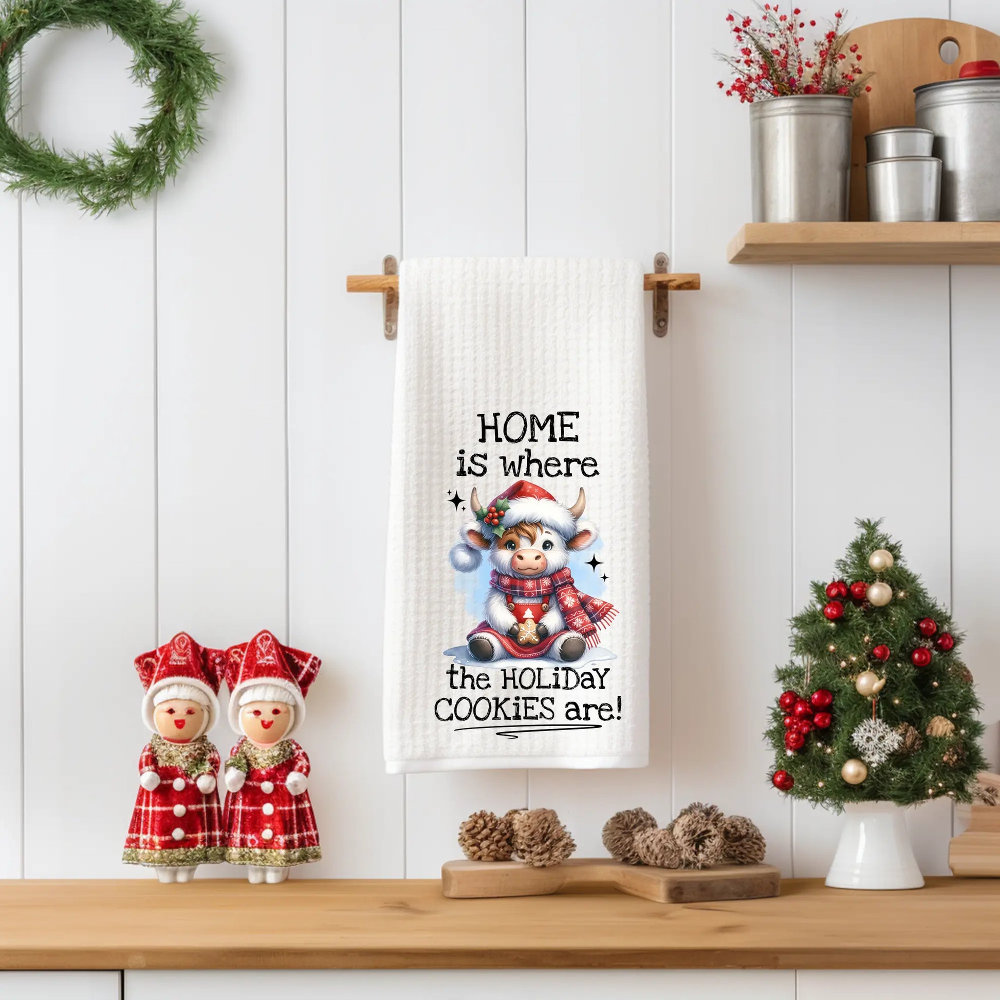 White 18x28 polyester waffle-weave tea towel hanging on a wooden peg rail, featuring a festive Highland cow in a Santa hat and plaid scarf holding a cookie with the text “Home Is Where the Holiday Cookies Are.” Displayed in a holiday kitchen scene with pinecones, a mini Christmas tree, and red-and-white figurines.