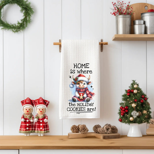 White 18x28 polyester waffle-weave tea towel hanging on a wooden peg rail, featuring a festive Highland cow in a Santa hat and plaid scarf holding a cookie with the text “Home Is Where the Holiday Cookies Are.” Displayed in a holiday kitchen scene with pinecones, a mini Christmas tree, and red-and-white figurines.