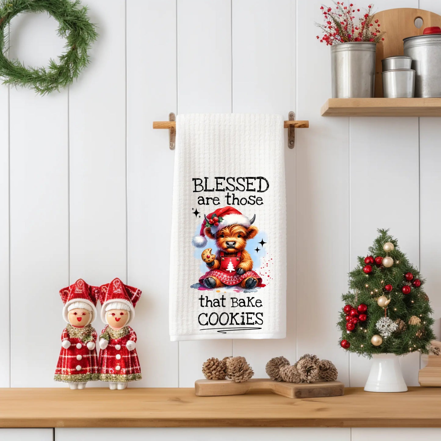 White 18x28 polyester waffle-weave tea towel hanging on a wooden peg rail, featuring a festive Highland cow in a Santa hat holding a cookie with the text “Blessed Are Those That Bake Cookies.” Displayed in a holiday kitchen scene with pinecones, a small Christmas tree, and red-and-white décor.