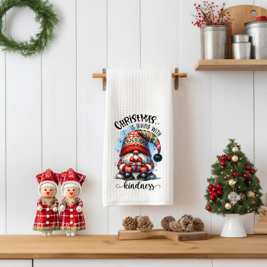 White 18x28 polyester waffle-weave tea towel hanging on a wooden peg rail, featuring a Christmas gnome with a patterned winter hat, mistletoe, and the text “Christmas Wishes & Mistletoe Kisses.” Displayed in a holiday kitchen scene with pinecones, a mini Christmas tree, and red-and-white décor.