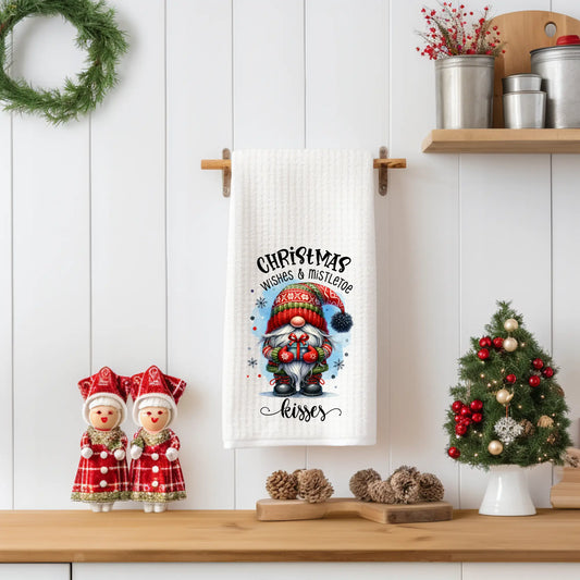 White 18x28 polyester waffle-weave tea towel hanging on a wooden peg rail, featuring a festive gnome holding a gift with the words “Christmas Is Giving With Kindness.” Displayed in a holiday kitchen scene with pinecones, a small decorated Christmas tree, and red-and-white decorative figurines.