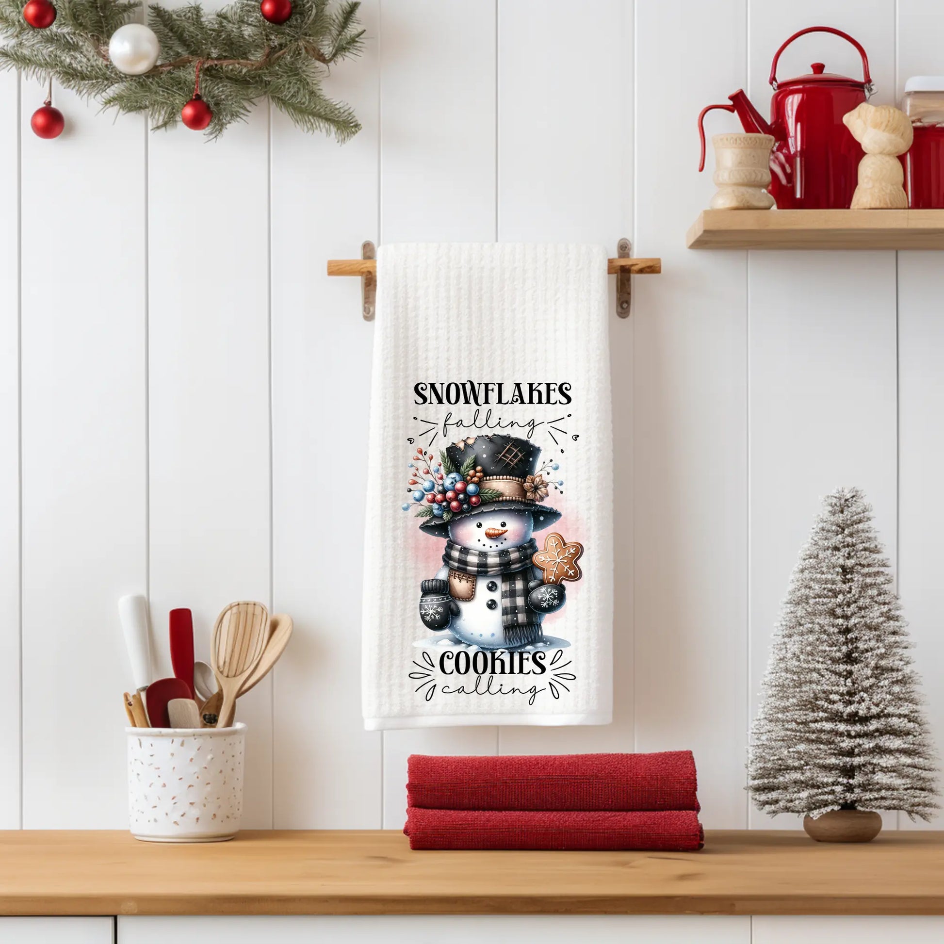 White 18x28 polyester waffle-weave tea towel hanging on a wooden peg rail, featuring a snowman with a plaid scarf, floral winter hat, and gingerbread cookie, along with the text “Snowflakes Falling, Cookies Calling.” Displayed in a festive kitchen scene with red towels, utensils, and a tabletop Christmas tree.