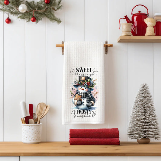 White 18x28 polyester waffle-weave tea towel hanging on a wooden peg rail, featuring a snowman with a plaid scarf, floral hat, and gingerbread cookie, with the text “Sweet Blessings, Frosty Nights.” Displayed in a holiday kitchen scene with red towels, a mini Christmas tree, and seasonal décor.