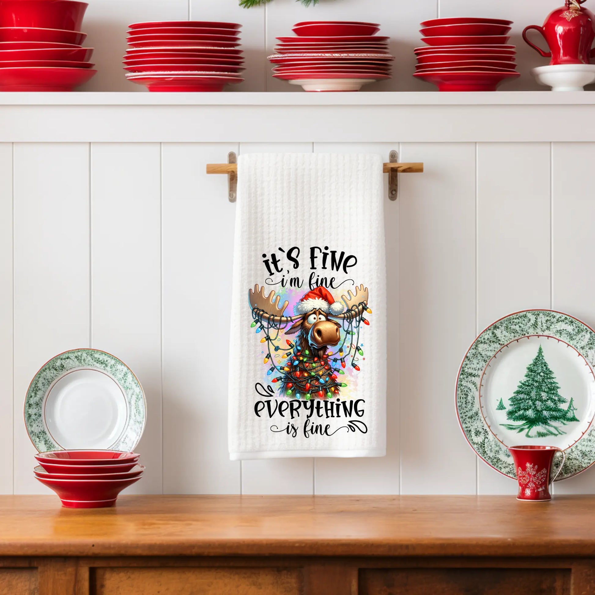 White 18x28 polyester waffle-weave Christmas tea towel hanging on a wooden peg rail, featuring a festive moose wearing a Santa hat and wrapped in colorful Christmas lights with the text “’Tis the Season to Be Jolly.” Displayed in a holiday kitchen scene with small Christmas trees, red teapots, and winter décor.