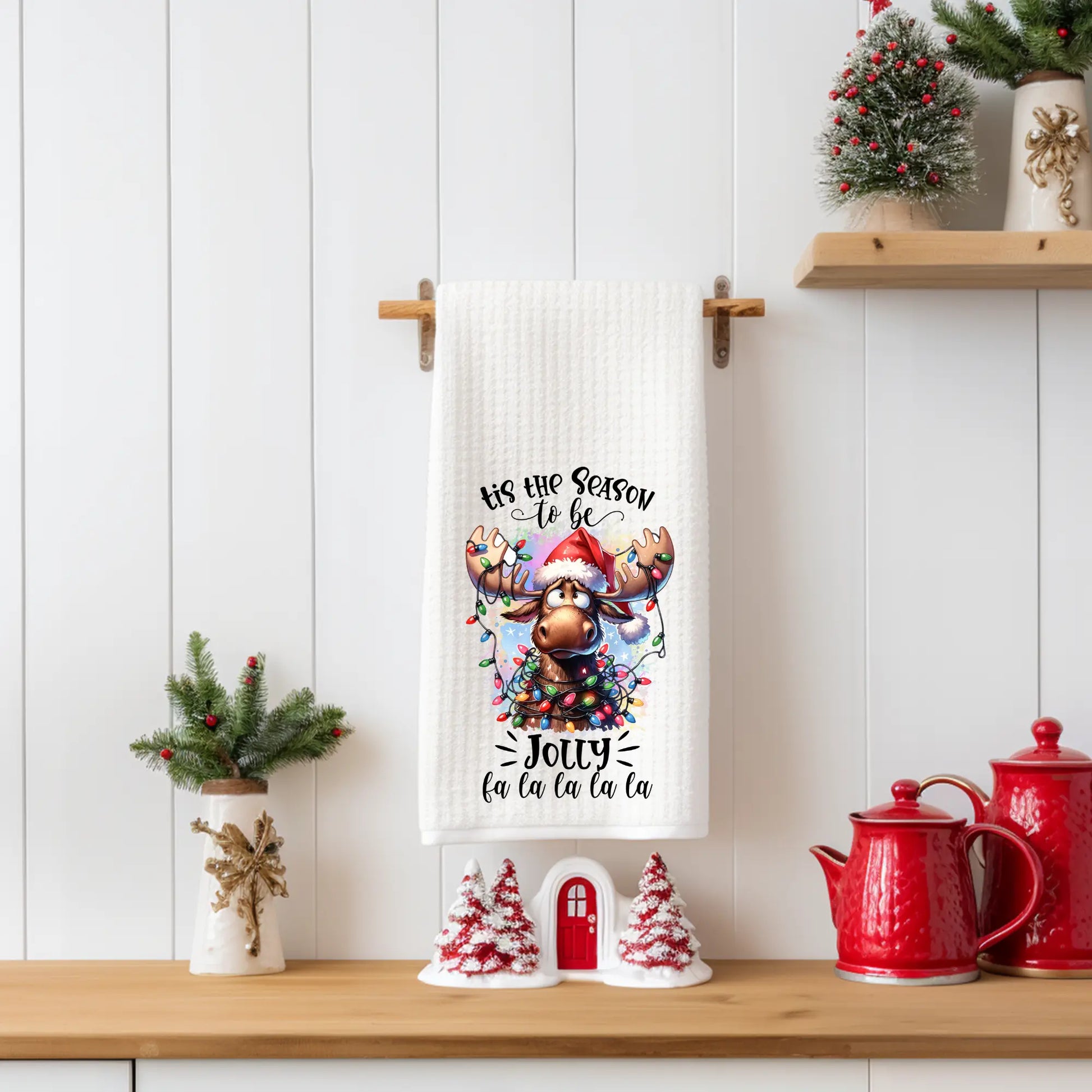 “Tis the Season to Be Jolly” Christmas tea towel featuring a cute moose wearing a Santa hat and wrapped in colorful Christmas lights, printed on a white 18x28 polyester waffle-weave towel.
