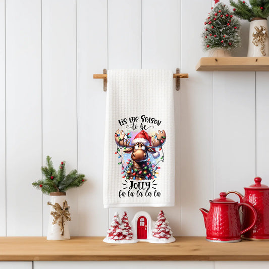 “Tis the Season to Be Jolly” Christmas tea towel featuring a cute moose wearing a Santa hat and wrapped in colorful Christmas lights, printed on a white 18x28 polyester waffle-weave towel.